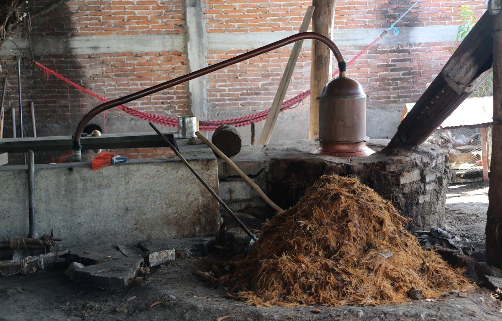 A copper pot still with its long arm, beside a pile of spent agave fiber